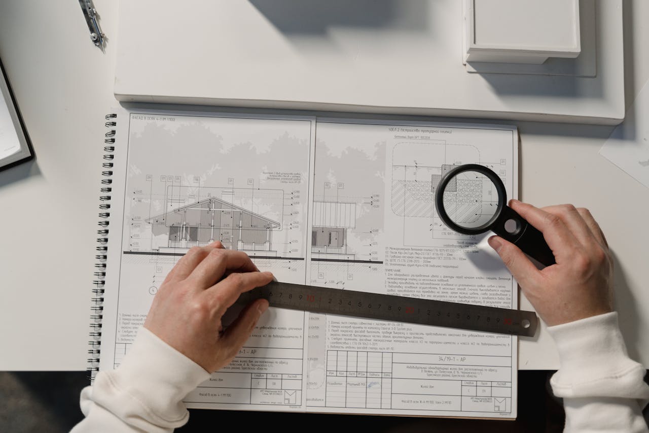 about-01 Hands holding a ruler and magnifying glass over architectural floor plans on a white table.